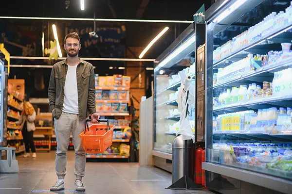 Man holding a shopping basket in a grocery store, standing next to refrigeration units, representing the need for reliable commercial appliance repair services in supermarkets.